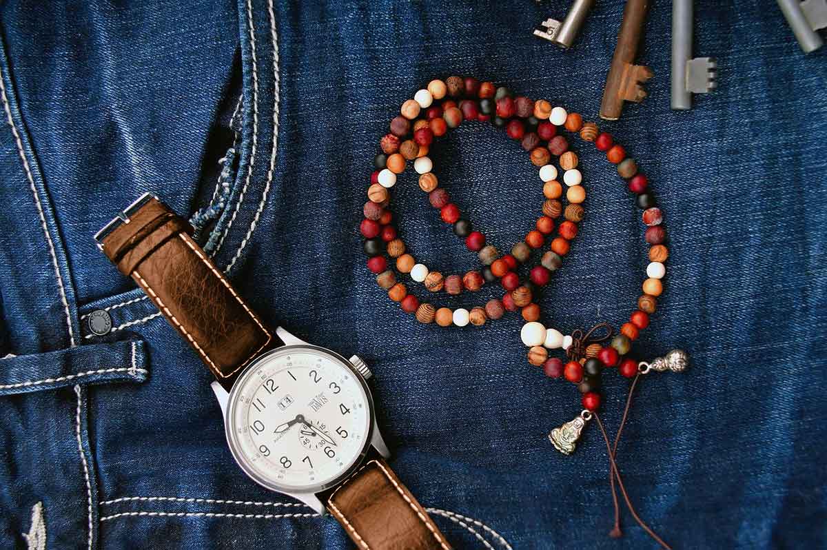 Braune Lederuhr und rotes, orangefarbenes und braunes Holzperlenarmband, das auf Blue Jeans liegt.