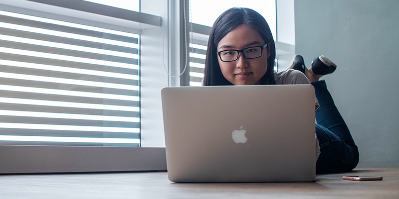 透明なガラス窓の横で銀色の MacBook を使用する 10 代の若者